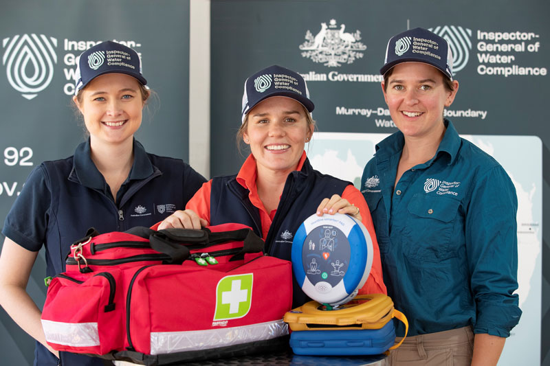 Three female IGWC employees standing around an AED and associated first aid kit.